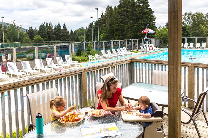 Une mère et ses deux enfants dinent sur la terrasse extérieure du centre récréatif. En arrière-plan, on aperçoit la piscine.