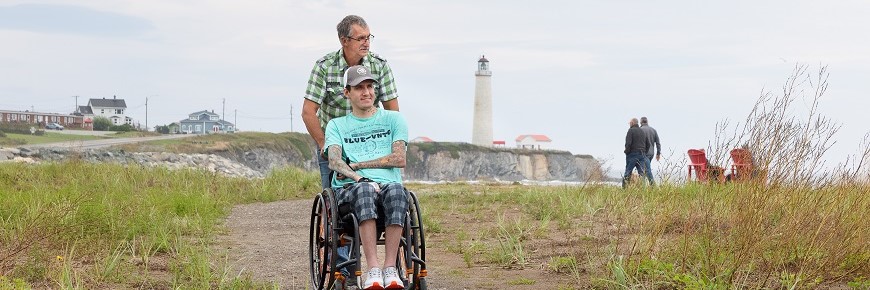 Un homme en pousse un autre assis dans un fauteuil roulant au sentier Du Banc. Derrière eux, on aperçoit le phare de Cap-des-Rosiers et des chaises rouges.