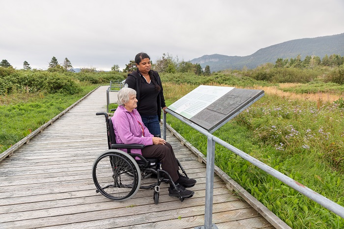 Deux femmes, dont une en fauteuil roulant, sont sur une passerelle de bois du sentier Prélude-à-Forillon et regardent un panneau d'interprétation traduit en braille. Il y a un garde-corps le long de la passerelle pour répondre au besoin d'une personne ayant un handicap visuel.