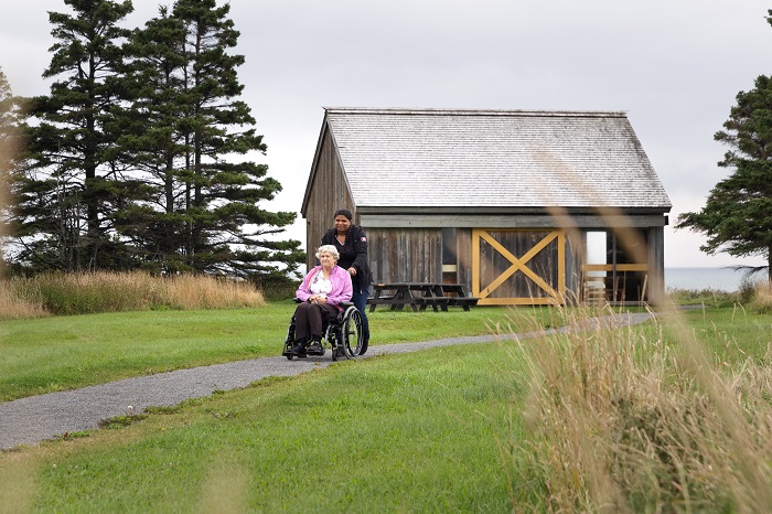 A woman pushes another in a wheelchair on the compacted stone dust portion of the Prelude-à-Forillon trail. A picnic building can be seen in the background.