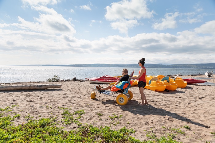 Une femme pousse un homme assis dans un fauteuil roulant tout-terrain sur le sable de la plage de Penouille. On aperçoit la baie de Gaspé en arrière-plan.