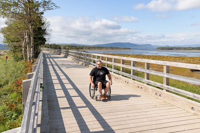 A man in a wheelchair heads along the wooden walkway of the Penouille trail. We see the salt marsh in the background.