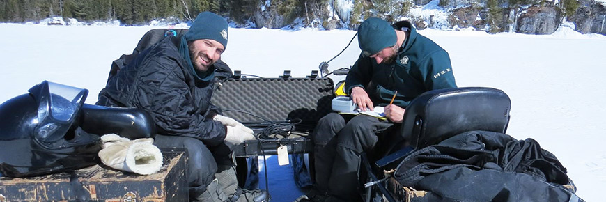 Sur un lac gelé, deux employés de Parcs Canada sont assis sur leur motoneige. L’un des employés prend des notes dans un cahier.