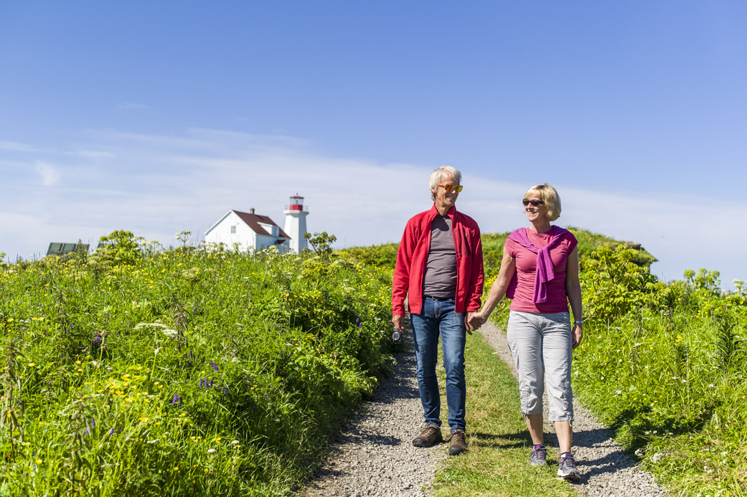 With the lighthouse keeper’s house and the lighthouse in the background, a couple is taking a stroll along the limestone trail on Île aux Perroquets