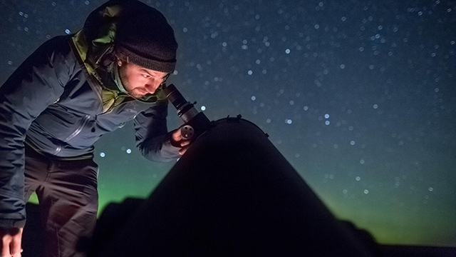 A man looks through a telescope at the starry night sky.