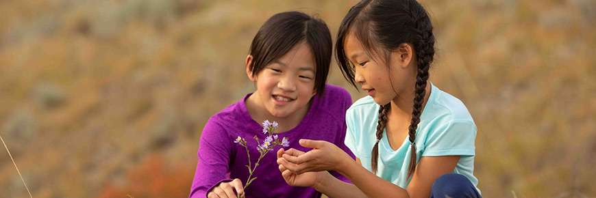 Two young girls crouch to examine a flower.