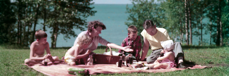 A family sitting on a blanket on a grassy lawn enjoys a picnic in front of a lake.