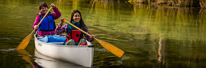 Two young ladies paddle a white canoe on a river. 