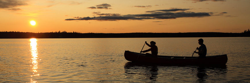 La silhouette d’un couple qui traverse un lac en canot se découpe sur un fond de coucher de soleil.
