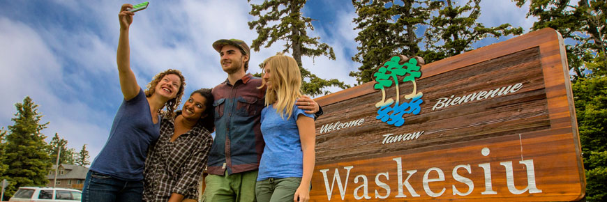 Four young adults take a ‘selfie’ in front of a sign welcoming visitors to the Town of Waskesiu.