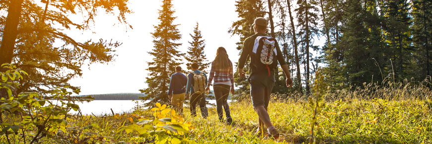 4 young adults hike through the forest toward a lake and the setting sun.