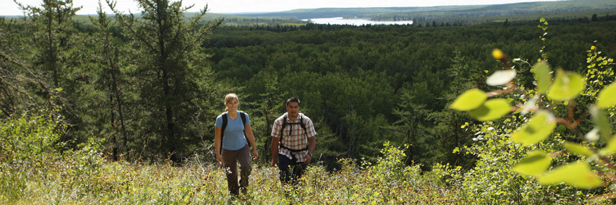 A young couple hike up a hillside with a scenic view of a forest lake behind them.