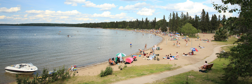 A motorboat is parked on the beach and there are many people enjoying various beach activities on the Waskesiu main beach. 