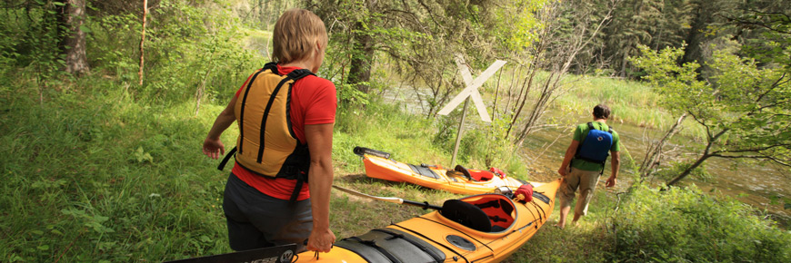 A man and a women carry a kayak to the river’s edge next to a portage marker. 