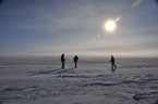 The shining sun hangs like a brilliant ball low in the blue sky above three people standing on flat snow covered ground that stretches off into the distance.