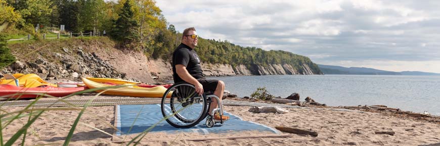 An adult in a wheelchair on a mobility mat on a sandy beach with kayaks, a pedal boat, and sandstone cliffs and in the background. 