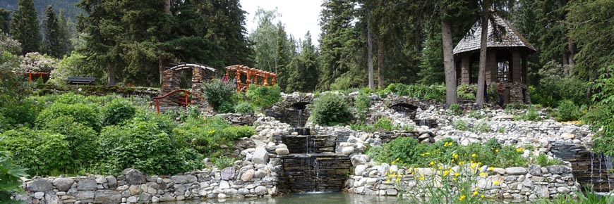 A stone waterfall and pond surrounded by greenery and small buildings in The Cascades of Time garden in Banff. 