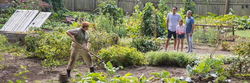 Une famille regarde un jardinier en costume d’époque qui s’occupe des nombreuses variétés de légumes d’un grand jardin. 
