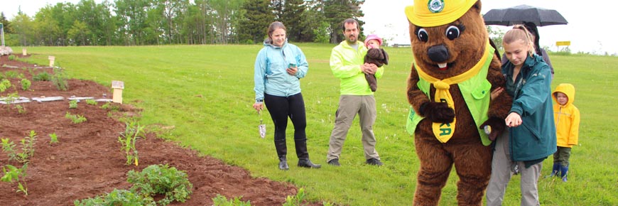 Un membre du personnel de Parcs Canada et la mascotte Parka font découvrir un jardin à une famille au parc national Fundy.
