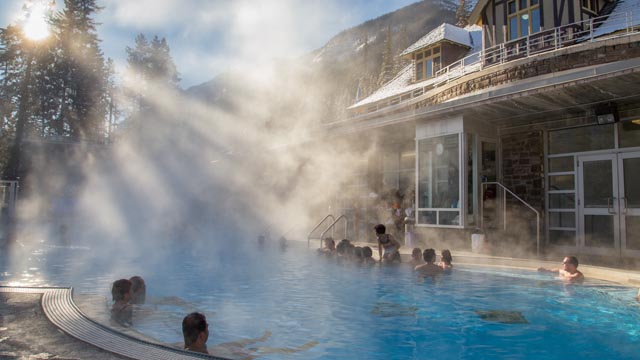 People enjoying a winter soak in Banff Upper Hot Springs with the sun shining at their backs. The historic bathhouse is in the background. 